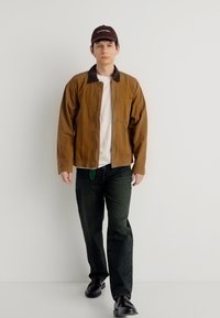 Νεαρός άνδρας φορώντας καφέ μπουφάν, λευκό πουκάμισο, μαύρο παντελόνι, μαύρα παπούτσια και σκούφο σκούρου καφέ, στέκεται μπροστά σε λευκό απλό τοίχο.