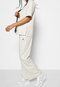 Personne portant un survêtement Jordan beige clair avec un pantalon à jambes larges et des baskets blanches, tenant la bandoulière d'un sac vert.