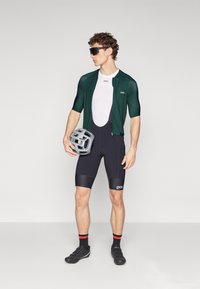 Cyklistický outfit pozostávajúci z krátkeho zeleného dresu s krátkym rukávom, čiernych šortiek s ramienkami a vystuženou textúrou a šedého prilby. Čierne cyklistické topánky a slnečné okuliare.