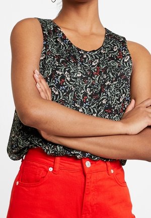 Kobieta z założonymi skrzyżowanymi ramionami, ubrana w czarny, bezrękawnik z kwiatowym wzorem i jasnoczerwone spodnie z wysokim stanem.