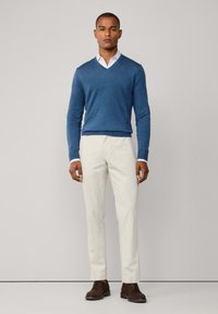 Maglione blu a V indossato sopra una camicia bianca, abbinato a pantaloni beige chiaro e scarpe stringate marroni. Tessuto morbido e vestibilità sartoriale.