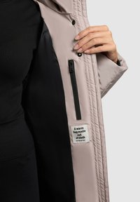 Persoon die een lichtbeige jas openhoudt met een zwarte voering, een binnentas met rits en een label waarop staat: "Een warme knuffel gemaakt van stof."