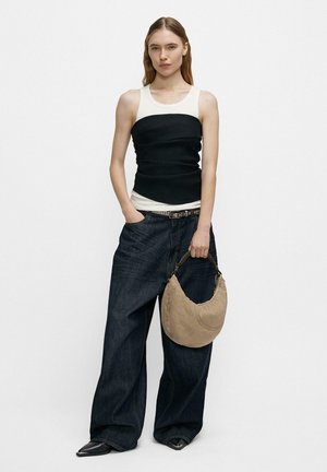 Frau trägt ein weißes und schwarzes ärmelloses Oberteil, weite dunkle Jeans, schwarze spitze Schuhe, hält eine beige Kuriertasche mit Kettenhenkel.