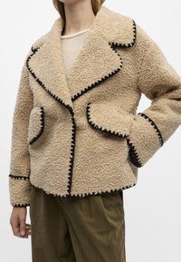 Beige Fleecejacke mit schwarzem Kontraststich, großen aufgesetzten Taschen und einem Schalkragen. Strukturierten Stoff mit lockerer Passform.