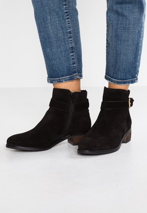 Bottines en daim noir avec fermeture éclair latérale, sangle et talon bas en bois, portées avec un jean bleu retroussé.