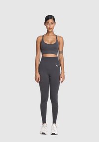 Žena stojí čelom dopredu, oblečená v tmavosivej športovej podprsenke a vysoko pásových legínach, bielych teniskách a s tetovaniami na rukách, na neutrálnom pozadí.