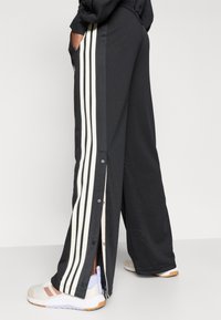 Čierne športové nohavice s bielymi bočnými pruhmi, so zapínaním na cvoky pozdĺž strán a sieťovinovou textúrou. Kombinované s bielymi teniskami.