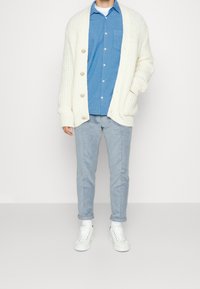 Cardigan a maglia crema con grandi bottoni sopra una camicia azzurra a bottoni, abbinato a pantaloni a righe chiare e sneaker bianche.