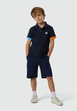 Garçon debout, les mains dans les poches, portant un polo bleu marine avec des manches à revers bleus et orange, un short cargo bleu marine, des chaussettes blanches et des baskets blanches.