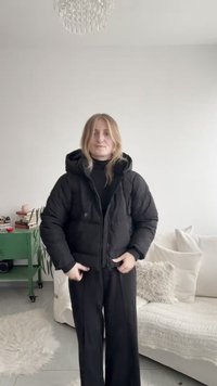 Black puffer jacket with a hood, featuring a zippered front and side pockets. Worn over a black turtleneck and paired with wide-leg pants.