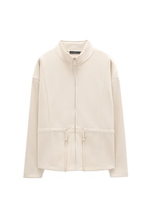 Beżowa kurtka na zamek z wysokim kołnierzem, regulowanymi sznureczkami w talii oraz długimi rękawami, przedstawiona na białym tle.