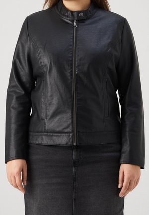 Person iført en sort jakke i kunstlæder med lynlås foran og trykknapkrave, kombineret med en mørkegrå denimnederdel, set fra taljen og op.