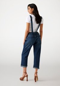 Denim-Latzhosen mit verstellbaren Trägern, dunkelblauer Farbe, lockerer Schnitt, umgeschlagener Saum und Gesäßtaschen. Kombiniert mit braunen Plateauschuhen.