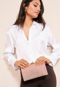 Ružičasta torbica od brušene kože sa zlatnim detaljima, koju drži osoba u bijeloj košulji s gumbima. Glatka tekstura, pravokutni oblik, suptilna elegancija.