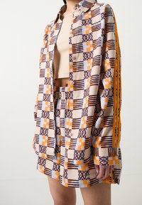 Camisa de manga corta con botones y estampado en beige, marrón y naranja, con diseños geométricos; complementada con pantalones cortos a juego y top color topo.