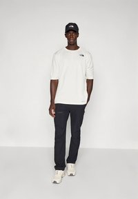 Valkoinen lyhythihainen t-paita, jossa on logo, tummat taskulliset housut ja beiget lenkkarit; asusteena musta lippalakki, jossa on logo.