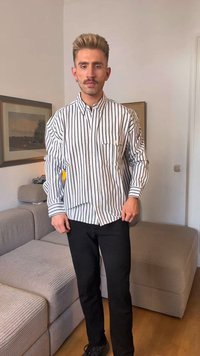Camicia a maniche lunghe a righe bianca e nera, con chiusura frontale a bottoni, colletto e taschino. Visibili pantaloni neri e divano beige texture.
