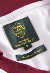 Officiële productlabel van AS Roma; groene stof met gele tekst, met een wolvenlogo, maat S, en een "Made in Europe" label op een bordeauxrode en witte shirt.