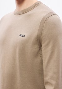 Beżowy sweter z długim rękawem z prążkowanym dekoltem i teksturowanymi akcentami na ramieniu. Logo "BOSS" w ciemnoniebieskim kolorze na klatce piersiowej.