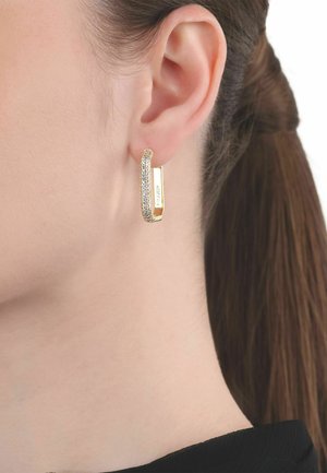 Primer plano de la oreja de una mujer con el cabello castaño liso recogido, que lleva un pendiente de aro rectangular dorado con pequeñas piedras incrustadas.