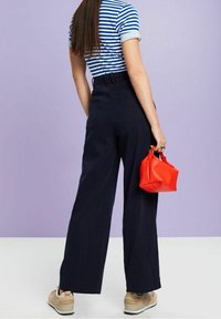 Marinblå vida byxor, randig blå och vit topp, håller en röd texturerad väska. Ljus bakgrund, informella skor synliga.