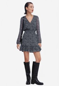 Bloemenprintjurk in zwart-wit, met lange doorschijnende mouwen, een V-hals, een gerimpelde zoom en een getailleerde taille. Gecombineerd met zwarte laarzen tot aan de knie.