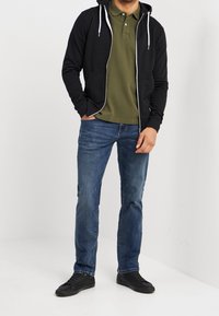 Černá mikina na zip s bílými tkaničkami, olivově zelené polo tričko, modré džínové kalhoty a černé tenisky. Neformální, vrstvený outfit.