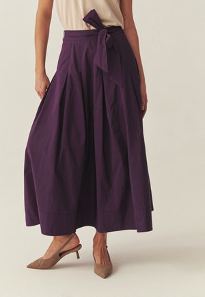 Žena oblečená v purpurovej plisovanej midi sukni s vysokým pásom a mašľou v páse, spolu s béžovými sandálmi na podpätku so zaťahovacím remienkom a špicatou špičkou.