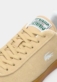 Μπεζ είδους σουέντ αθλητικό παπούτσι με υφασμάτινα κορδόνια, λαστιχένια σόλα και λευκή ετικέτα στη γλώσσα με πράσινο κείμενο. Εμφανισμένο ανάγλυφα το λογότυπο της Lacoste στο πλάι.