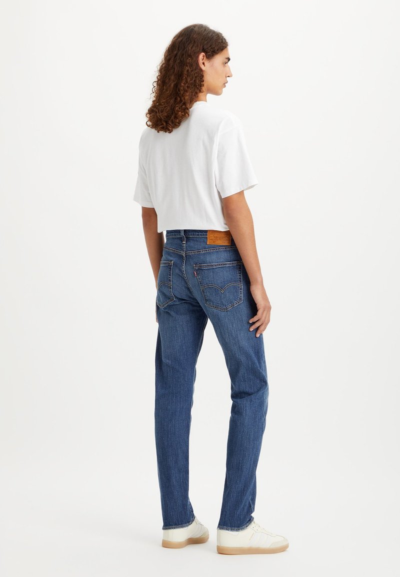 Blaue Jeans mit geradem Bein, ausgestattet mit einem beigefarbenen Lederpatch auf der Rückseite, kombiniert mit einem weißen, verkürzten T-Shirt und weißen Sneakers.