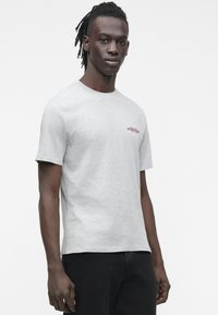 A szürke pamut póló kerek nyakkivágással és rövid ujjakkal rendelkezik, a mellrészen egy kis piros logóval. Az anyag sima tapintású.