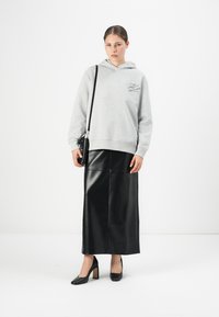 Donna che indossa un hoodie grigio, una gonna in pelle nera lunga fino alla caviglia, tacchi alti neri, e tiene una borsa a spalla nera, in piedi su uno sfondo bianco.