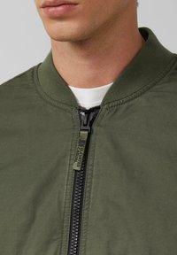 Olivgrüne Bomberjacke mit geripptem Kragen, Reißverschluss und einem Label am Reißverschluss. Das Material wirkt strukturiert und strapazierfähig.