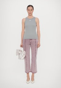Graues ärmelloses Tanktop, hellrosa ausgestellte Jeans mit rohem Saum, weiße Absatzschuhe, hält eine glänzende silberne Tasche mit einem Schleifendetail.