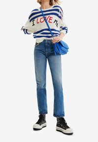Suéter blanco de punto con rayas azules y rojas, que presenta el texto "ME AMO". Combinado con jeans azul claro y zapatillas de altura negra con estampado.