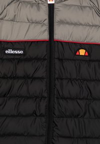 Czarna i szara pikowana kurtka z zamkiem z przodu, czerwoną akcentującą taśmą oraz haftowanym logo na piersi z okrągłym graficznym detalem.