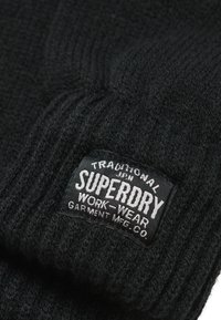 Gorro de punto negro con canalé texturizado, que presenta una etiqueta rectangular negra con texto blanco que dice "SUPERDRY WORK-WEAR."