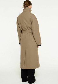 Lång beige ullkappa med hög krage och bälte i midjan. Har vida ärmar och rak skärning, som sträcker sig till mitten av vadden med ett sprund bak.