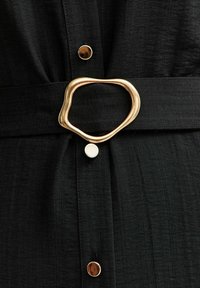 Indumento in tessuto nero con superficie testurizzata, una cintura sottile con una fibbia d'oro astratta e bottoni metallici a contrasto.