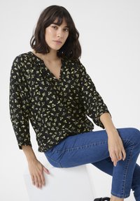 Blouse noire à motifs floraux jaunes, décolleté en V et manches trois-quarts. Associée à un jean bleu et assise sur un cube blanc.