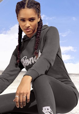 Dunkelgrauer Kapuzenpullover mit "Bench"-Logo, kombiniert mit passenden Leggings. Geflochtenes Haar, minimale Accessoires, glatte Stoffstruktur.