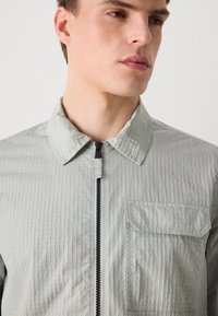 Ανοιχτό πράσινο σακάκι με γιακά, με υφή στην επιφάνεια, πλήρη φερμουάρ και μπρο�στινή τσέπη. Διαθέτει μια διακριτική σχεδίαση με καρέ.