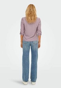 Maglietta lunga viola chiaro con maniche arrotolate, abbinata a jeans azzurri a gamba larga con tasche posteriori e leggero sfilacciamento sui bordi.