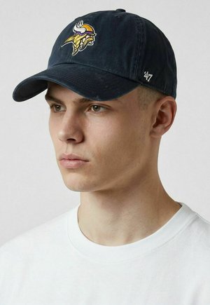 Junger Mann trägt eine dunkelblaue Baseballkappe mit gelbem Wikinger-Logo und ein weißes T-Shirt mit Rundhalsausschnitt, schaut seitlich vor einem schlichten Hintergrund.