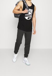 Homme portant un haut noir sans manches Puma, un pantalon de jogging foncé, des baskets blanches, avec un sac à dos à motif porté sur une épaule, sur un fond uni.