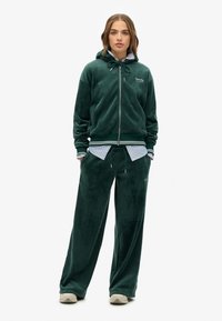 Tuta verde vellutato con una felpa con zip e pantaloni con cordino. Polsini e orlo a costine, indossato sopra una maglietta a righe blu, con dettagli del logo.