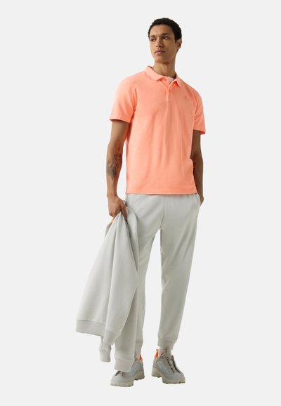 Muž v korálovém polokošili a světle šedých kalhotách drží odpovídající sako, má na sobě šedé tenisky s oranžovými detaily, stojí před bílým pozadím.