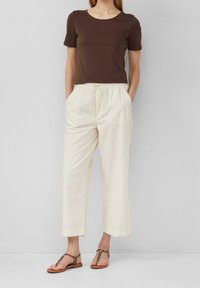 Brązowa koszula z krótkim rękawem, kremowe spodnie z elastycznym pasem oraz sandały w zwierzęcy wzór. Spodnie mają luźny krój, boczne kieszenie i proste nogawki.