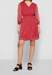 Mujer con un vestido rojo de flores hasta la rodilla, mangas largas transparentes, tacones negros abiertos en la punta y sosteniendo un pequeño bolso de cadena.