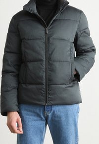 Giacca a vento nera con texture trapuntata, colletto alto e zip frontale, abbinata a jeans in denim azzurro chiaro. Dettagli notevoli includono tasche laterali.
